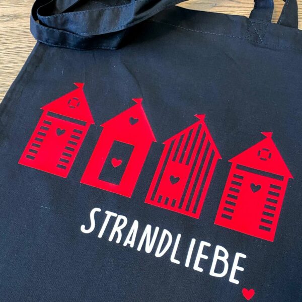 Ausschnitt Baumwolltasche schwarz mit weissem Aufdruck "Strandliebe" und roten Strandhäuschen