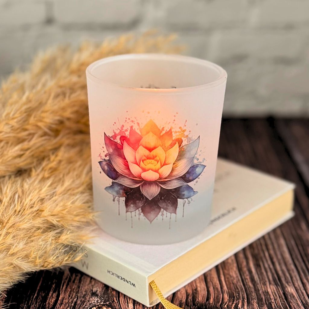 Satiniertes Teelichtglas beleuchtet mit bunter Lotusblüte im Watercolor-Stil auf einem Buch platziert.