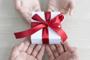 Titelbild Blogartikel zum Thema Schenken: Hände überreichen ein weiss eingepacktes Geschenk mit roter Schleife